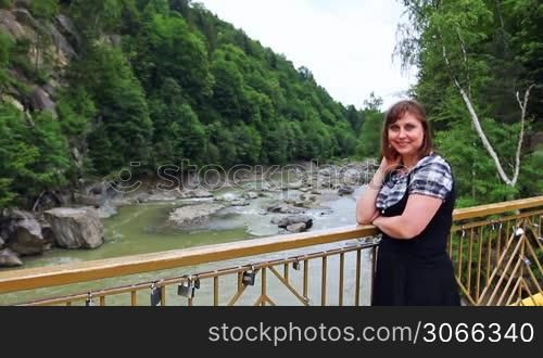 pretty woman stands near railing bridge, then looks on river in Carpathian Mountains