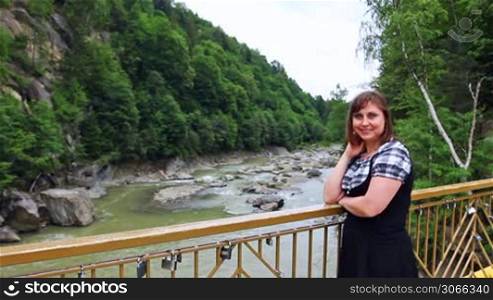 pretty woman stands near railing bridge, then looks on river in Carpathian Mountains