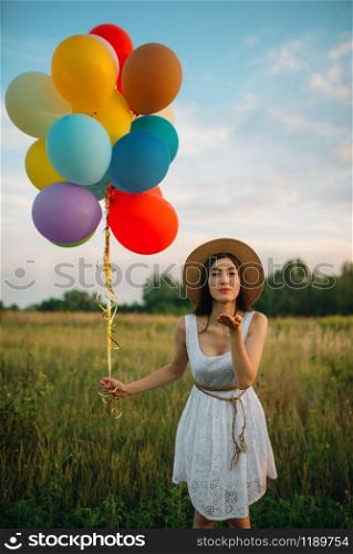 Pretty woman with bunch of colorful balloons sends a kiss, green field on background. Cute girl on summer meadow at sunny day