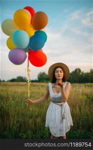 Pretty woman with bunch of colorful balloons sends a kiss, green field on background. Cute girl on summer meadow at sunny day