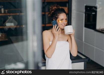 Pretty woman with cosmetic patches under eyes drinks morning coffee talking via smartphone at home. Young female wrapped in towel answering call by phone during beauty skincare treatment.. Pretty girl with under eyes patches drinks morning coffee talking by phone during skincare at home