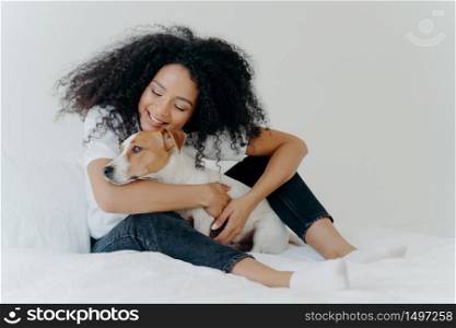 Pretty woman with curly hairstyle plays and enjoys with adorable small dog in bed, has good mood, pose together in cozy bedroom. Female wakes up with best friend. Lovely pet with owner indoor