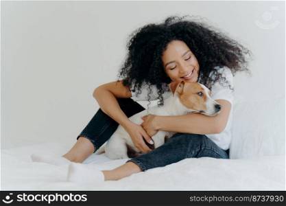 Pretty woman with curly hairstyle plays and enjoys with adorable small dog in bed, has good mood, pose together in cozy bedroom. Female wakes up with best friend. Lovely pet with owner indoor