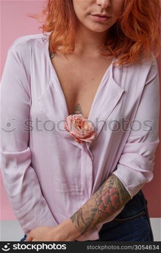 Pretty woman with ginger hair, tattoo on her bust and hands holds fresh rose living coral color on a light background. . Sexual girl with fresh flower living coral color in hands with tattoo on a gray background.