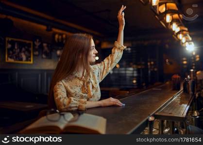 Pretty woman with raised hand sitting at the counter in bar. One female person in pub, human emotions, leisure activities, nightlife. Woman with raised hand sitting at counter in bar
