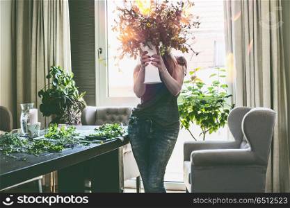 Pretty women holding vase with summer bouquet of wild flowers in her hands in living room with table and ears armchair at window. Home lifestyle and decor ideas