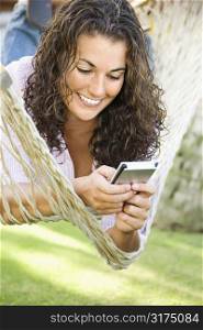 Pretty young adult Caucasian brunette female lying in hammock using PDA and smiling.