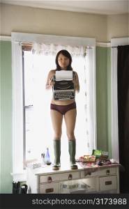 Pretty young Asian woman in underwear and rubber boots holding typewriter.