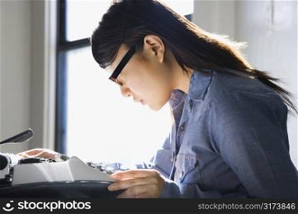 Pretty young Asian woman sitting in chair typing on typewriter in kitchen.