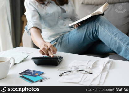 Pretty young Asian woman using calculator while holding notebook to note home expenses and taxes in living room at home.