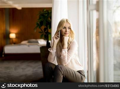 Pretty young blonde woman with mobile phone by the window in the room