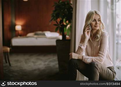 Pretty young blonde woman with mobile phone by the window in the room