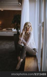 Pretty young blonde woman with mobile phone sitting by the window in the room