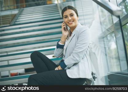 Pretty young business woman sitting on the stairs at the office and use mobile phone