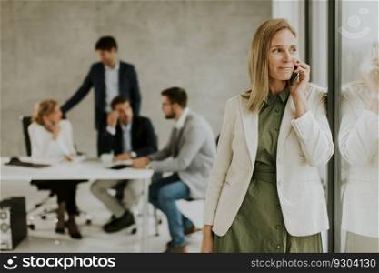 Pretty young business woman standing in the office and using mobile phone in front of her team