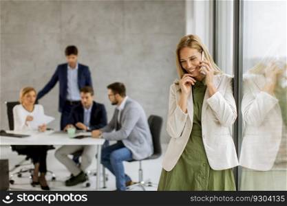 Pretty young business woman standing in the office and using mobile phone in front of her team