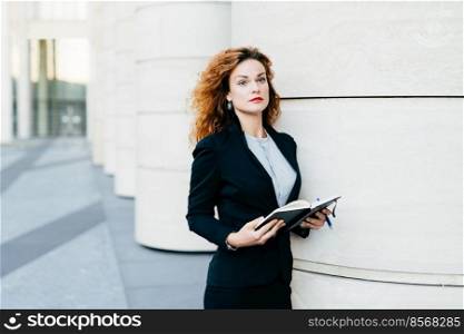 Pretty young businesswoman wearing black jacket, skirt and white blouse, holding her pocket book with pen, writing notes or looking for spare days in order to arrange meeting with companions