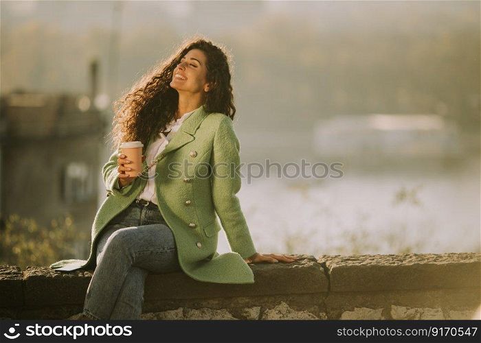 Pretty young curly hair woman enjoying autumn sun while sitting by the river and drinking takeaway coffee