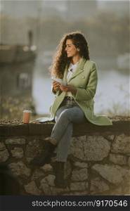 Pretty young curly hair woman using smartphone while sitting by the river and drinking takeaway coffee