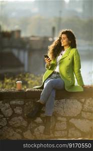 Pretty young curly hair woman using smartphone while sitting by the river and drinking takeaway coffee