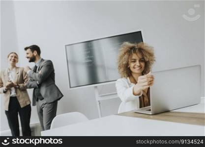 Pretty young curly hair woman working on laptop in bright office with team member discussing behind her