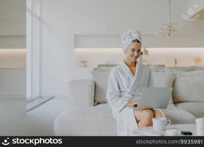 Pretty young European woman dressed in comfortable domestic clothes works on laptop computer at home sits on sofa against modern apartment interior wears minimal makeup smiles toothily at camera