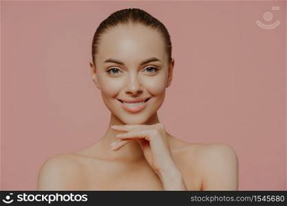 Pretty young European woman touches chin gently, enjoys flawless of skin after beauty procedures, poses nude, has natural makeup, isolated on pink background. Beauty, spa and wellness concept