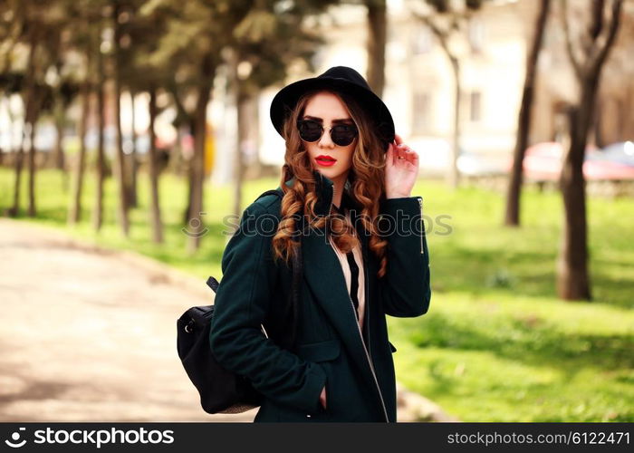 Pretty young fashion smiling blonde woman in sunglasses and vintage hat with leather bag posing outdoor in green park background in spring sunny weather. Fashion image of stylish hipster girl