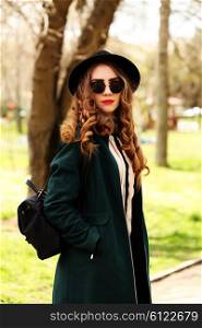 Pretty young fashion smiling blonde woman in sunglasses and vintage hat with leather bag posing outdoor in green park background in spring sunny weather. Fashion image of stylish hipster girl