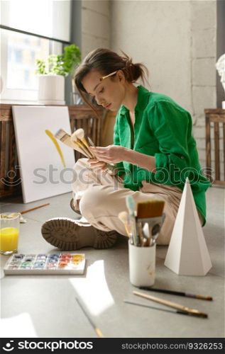 Pretty young female artist drawing picture on canvas with oil paints sitting on floor in her own art studio. Creativity in work and contemporary creative painter concept. Young female artist drawing picture sitting on floor in her own art studio