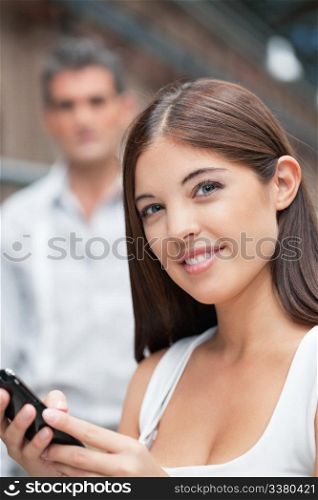 Pretty young female holding cell phone with man in background