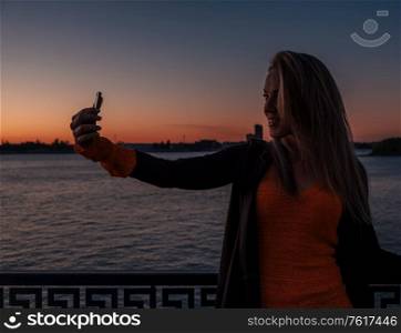 Pretty young female in knitted sweater holding takes selfie while on city street at night in front of river sunset. Pretty female holding takes selfie while on city street at night in front of river sunset