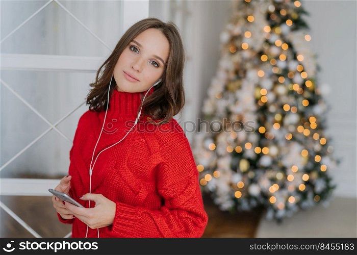 Pretty young female model listens pleasant music in earphones, holds cellular, dressed in red knitted sweater, poses in living room near Christmas tree with blank space for your advertising content