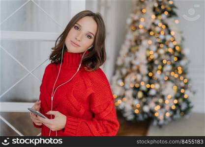 Pretty young female model listens pleasant music in earphones, holds cellular, dressed in red knitted sweater, poses in living room near Christmas tree with blank space for your advertising content