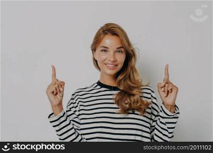 Pretty young female with dyed blonde hair pleasantly smiling while gesturing pointing with index fingers upwards at copy space, shows something above her head, isolated over grey background in studio. Young female with dyed blonde hair pleasantly smiling while pointing with index fingers upwards