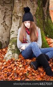 Pretty young girl sitting beside a tree in Autumn with a carpet if leaves on the floor