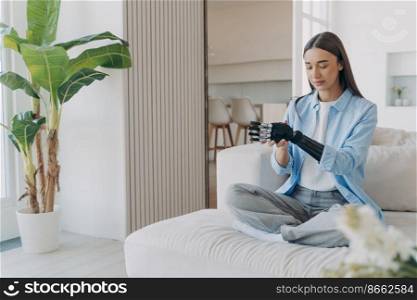 Pretty young girl with disability installing her bionic prosthetic arm, sitting on sofa in modern living room at home. Disabled woman using artificial limb, high tech robotic hand.. Girl with disability uses bionic prosthetic arm, sitting on sofa in modern living room at home