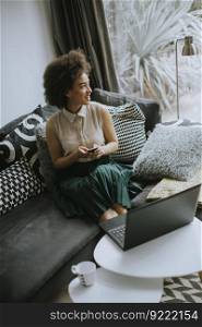 Pretty young lady with curly hair work on the notebook while sit down on the couch at home