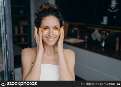 Pretty young lady wrapped in towel after bathing is happy and relaxing at home or hotel room. European girl applying eye patches. Spa resort, body care. Having rest on weekend or vacation concept.. Pretty young lady wrapped in towel after bathing is happy and relaxing applying eye patches.