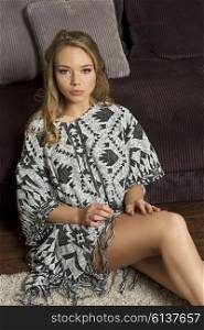 pretty young model , sitting on the floor near a sofa , she is wearing casual dress , and she is posing looking at the camera.