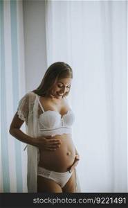 Pretty young pregnant woman standing at window in the room at daylight