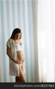 Pretty young pregnant woman standing by the blue stripped wall in the room at daylight