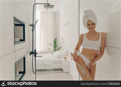 Pretty young woman dressed in white underwear doing dry leg massage with wooden organic brush after taking morning shower in bathroom at modern apartment interior. Women beauty and body care concept. Pretty young woman doing dry leg massage with wooden organic brush