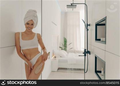 Pretty young woman dressed in white underwear doing dry leg massage with wooden organic brush after taking morning shower in bathroom at modern apartment interior. Women beauty and body care concept. Pretty young woman doing dry leg massage with wooden organic brush