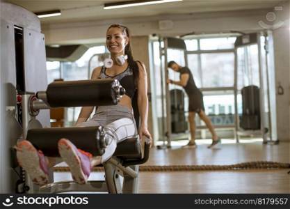 Pretty young woman having exercises on leg extension and leg curl machine in the gym