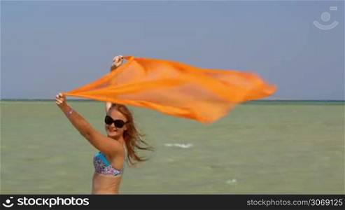 Pretty young woman holding pareo on the beach on a windy day and looking to the camera with smile