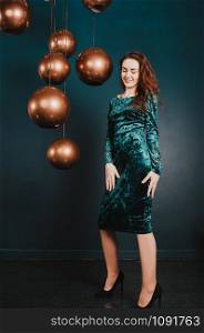 Pretty young woman in a green blue velvet dress, posing next to hanging big golden balls, long legs, high heels, dark indigo backdrop. New Year, Christmas holiday