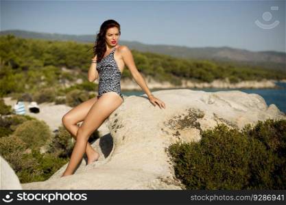Pretty young woman in bikini sitting on beach rocks by the sea