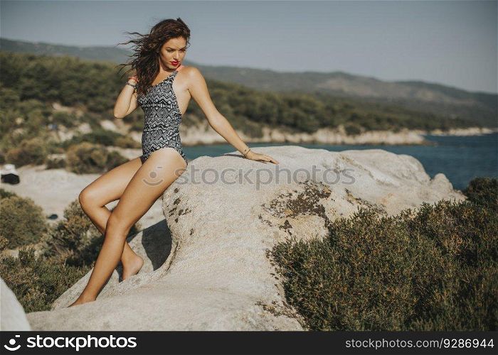 Pretty young woman in bikini sitting on beach rocks by the sea