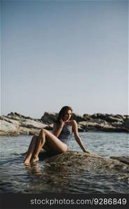 Pretty young woman in bikini sitting on rock by the sea